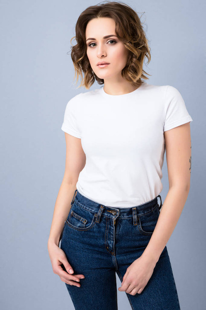 Portrait of stylish young girl in basic white t-shirt and high waisted blue jeans  posing in studio on grey background - Фото, изображение