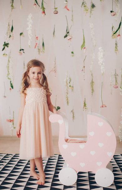 Schattig klein meisje in jurk met baby wandelwagen thuis, gelukkige jeugd concept  - Foto, afbeelding