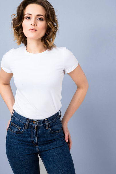 Portrait of stylish young girl in basic white t-shirt and high waisted blue jeans  posing in studio on grey background - Foto, imagen