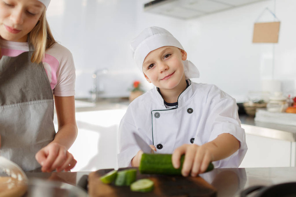 Un niño guapo cocinando en la cocina. Un niño corta pepinos frescos en una tabla de madera. Preparación de platos. El concepto de un alimento saludable
. - Foto, imagen