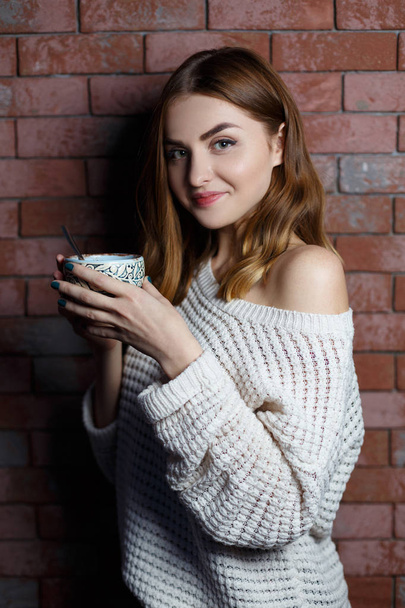 Jonge mooie vrouw, drinken een kopje koffie in het café op een rode bakstenen muur achtergrond. Stijlvolle dame met warme dranken. - Foto, afbeelding