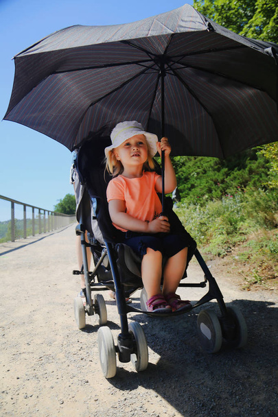 Bebek arabası içinde oturan küçük kız. Güneş'ten büyük şemsiye tarafından korunan çocuk. Yaz aylarında güneşli çocuk sıcak çarpması önleme - Fotoğraf, Görsel