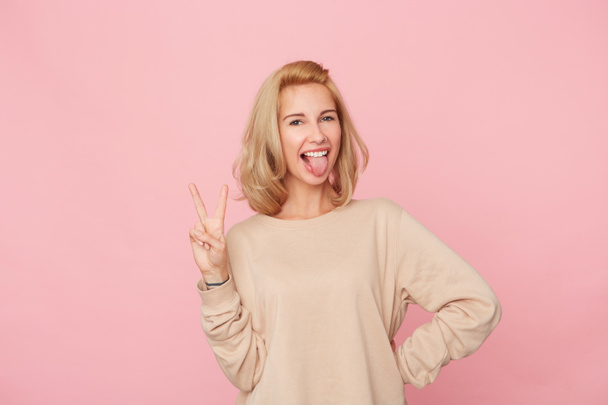 fröhliche blonde Frau, die vor rosa Hintergrund steht, lächelt und mit ihren Fingern und ihrer Zunge V-Zeichen zeigt. Porträt einer glücklichen Frau. Positives Konzept - Foto, Bild
