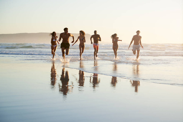 Group Of Friends Run Through Waves Together On Beach Vacation - Valokuva, kuva