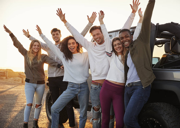 Young adult friends on a road trip standing by their jeep raising hands  - Фото, изображение
