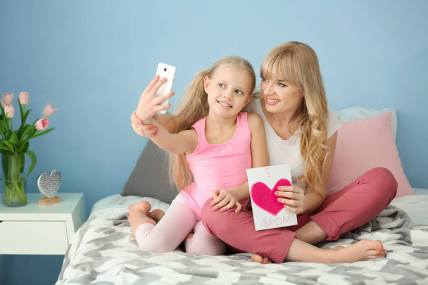 Linda niña y su madre tomando selfie con tarjeta hecha a mano en el dormitorio
 - Foto, Imagen
