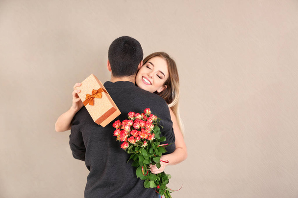 Ευτυχισμένος νεαρή γυναίκα με το κιβώτιο δώρων και λουλουδιών που αγκαλιάζει το φίλο της στο χρώμα φόντου - Φωτογραφία, εικόνα
