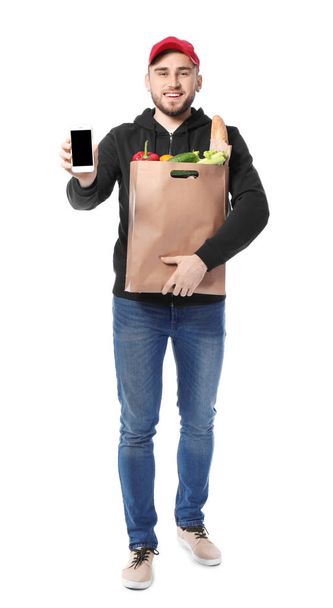 Young man with products in paper bag and mobile phone on white background. Food delivery service - Valokuva, kuva
