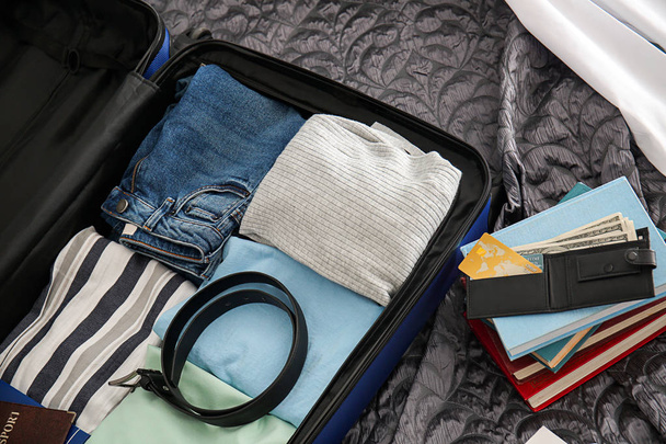 Brieftasche, Bücherstapel und offener Koffer mit gepackten Sachen auf dem Bett - Foto, Bild