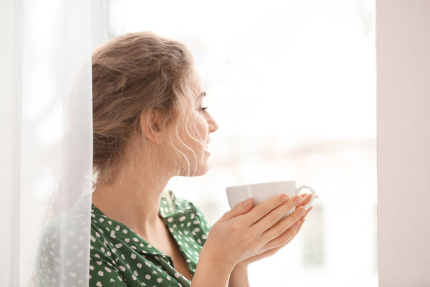 Young woman drinking tea near window indoors - Foto, Imagen