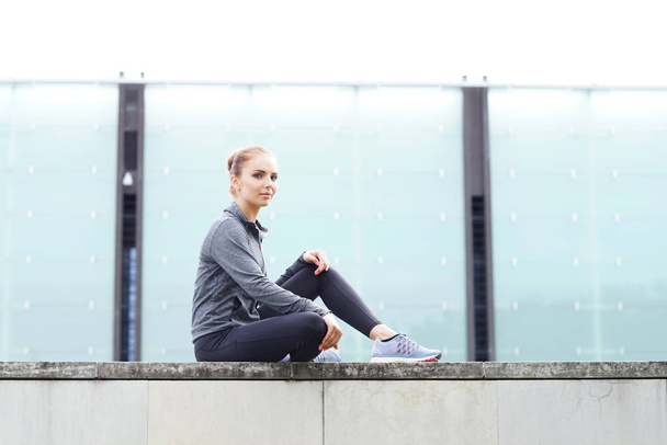Young, fit and sporty girl sitting on a concrete border. Fitness, sport, urban jogging and healthy lifestyle concept. - Foto, Imagen
