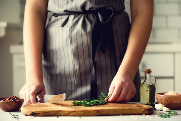 Woman hands cutting fresh green rosemary on wood chopping board in white kitchen, interior. Copy space. Homemade food conceplt, healthy recipe. Take me to work - Zdjęcie, obraz