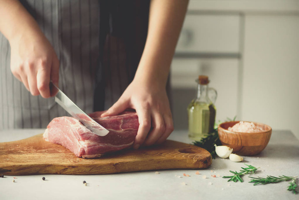 Wooman hands cutting beef meat on wooden chopping board, rosemary, oil, salt, pepper. Girk cooking pork meat on white kitchen backgound. Copy space - Photo, image