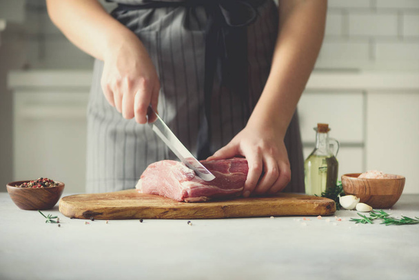 Wooman hands cutting beef meat on wooden chopping board, rosemary, oil, salt, pepper. Girk cooking pork meat on white kitchen backgound. Copy space - Foto, afbeelding