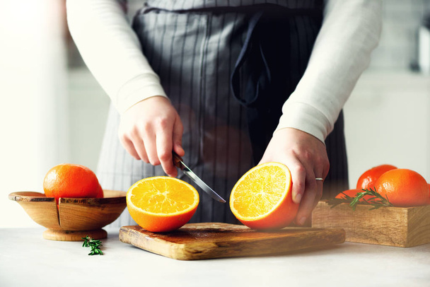 Woman hands slicing orange, cutting citrus fruit. Knife, wooden cutting board on design white kitchen. Copy space - Photo, Image