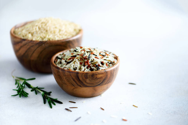 Raw organic brown rice in wooden bowl and rosemary on light concrete background. Food ingredients. Copy space - Foto, imagen