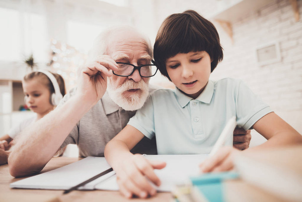 Grandpa helping boy with homework while girl listening music  - 写真・画像