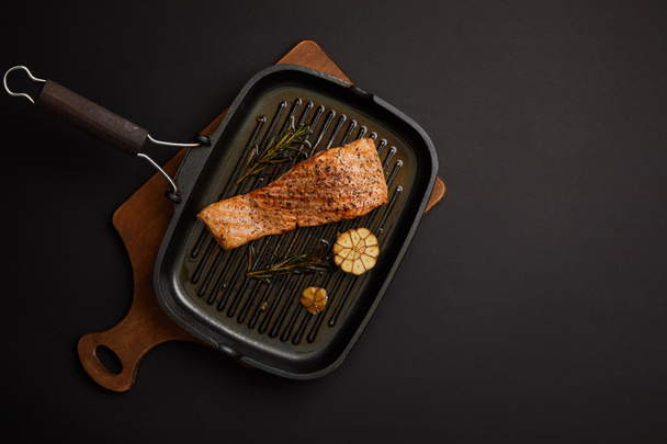 top view of arranged grilled salmon steak with rosemary on wooden cutting board on black tabletop - Фото, изображение