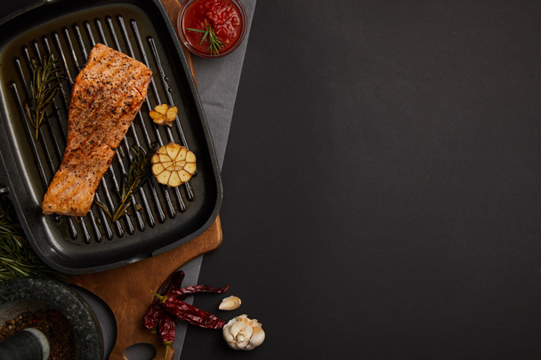 top view of arranged grilled salmon steak on wooden cutting board, ingredients and sauce on black tabletop - 写真・画像