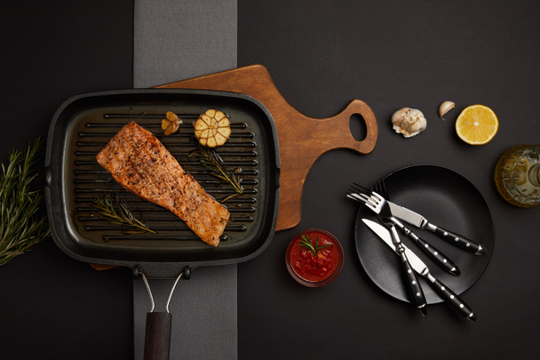 top view of arranged grilled salmon steak on wooden cutting board, sauce and cutlery on black tabletop - Photo, Image