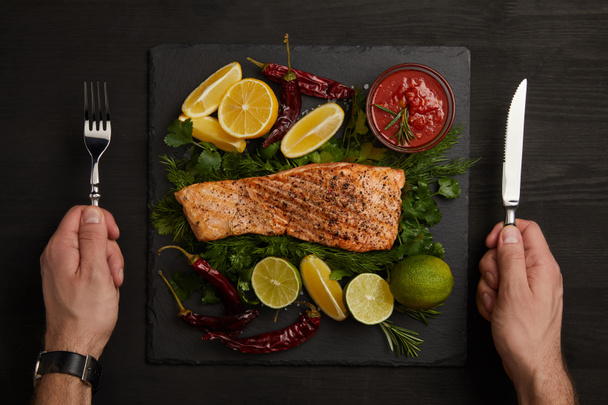 partial view of male hands with cutlery, grilled salmon steak, sauce and arranged citrus fruits on black surface - Photo, Image