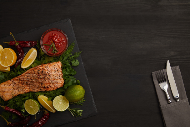 top view of grilled salmon steak, pieces of lime and lemon, sauce and cutlery on black surface - Valokuva, kuva