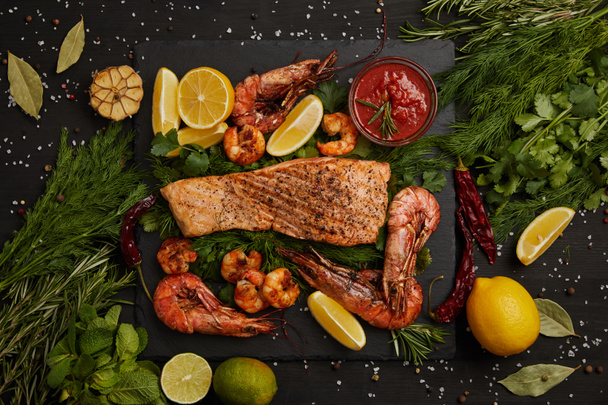 top view of grilled salmon steak, shrimps, pieces of lemon, sauce and spices on black surface - Photo, Image