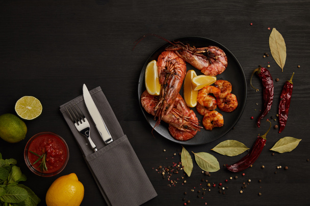 flat lay with grilled shrimps with lemon pieces on plate and arranged sauce, spices, ingredients and cutlery around on black surface - Photo, Image