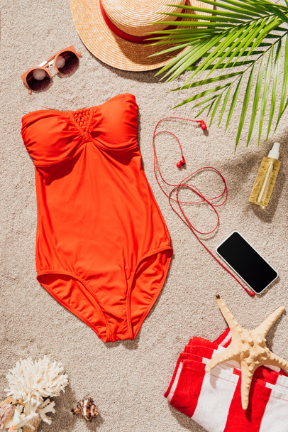 top view of stylish red swimsuit with smartphone and accessories lying on sandy beach - Photo, Image