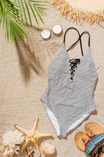 top view of stylish swimsuit with sunglasses and flip flops lying on sandy beach - Foto, afbeelding