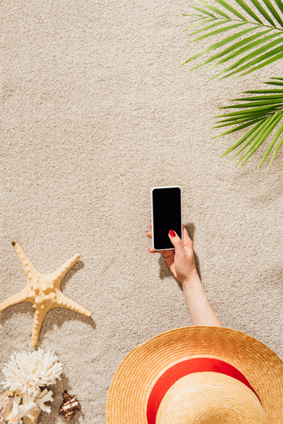 cropped shot of woman in hat using smartphone while lying on sandy beach - Zdjęcie, obraz