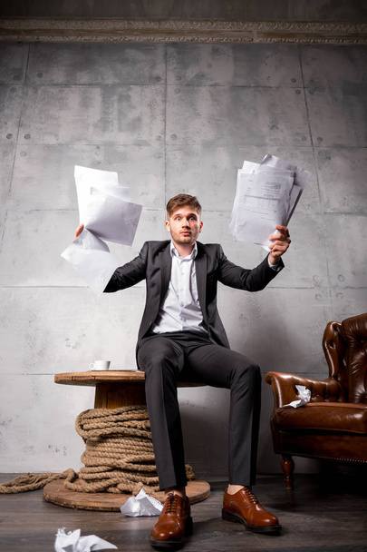 junger wütender Geschäftsmann sitzt auf Holztisch und wirft Papiere weg - Foto, Bild