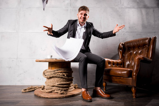 Young angry businessman sitting on wooden table holding documents in hands, full length - Фото, зображення