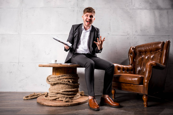 Young angry businessman in suit  sitting on wooden table in modern office holding documents and shouting  - Foto, Bild