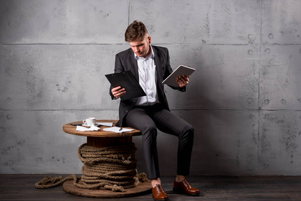 Молодой бизнесмен в костюме сидит на деревянном столе в офисе с цифровыми планшетами и документами
 - Фото, изображение