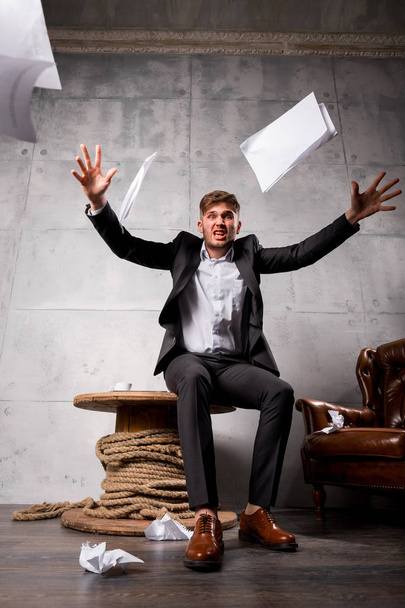 Jonge boos zakenman zittend op een houten tafel en papieren weg te gooien - Foto, afbeelding