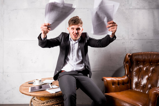 junger wütender Geschäftsmann sitzt auf Holztisch und wirft Papiere weg - Foto, Bild