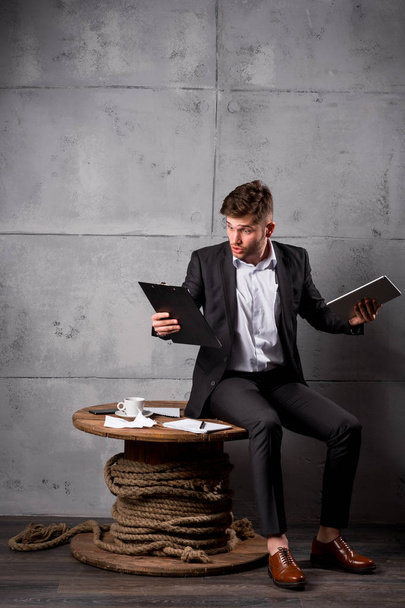 Young businessman in suit sitting on wooden table in office holding digital tablet and documents - Foto, Imagen