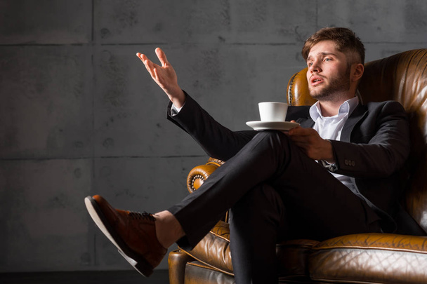 Молодой бизнесмен в костюме сидит на кожаном диване, держа чашку кофе
 - Фото, изображение
