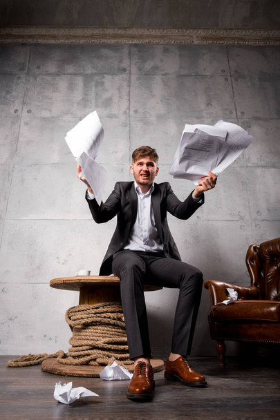 Young angry businessman sitting on wooden table and throwing away papers - Photo, Image
