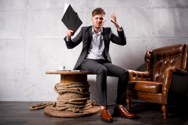 Young angry businessman in suit  sitting on wooden table in modern office holding documents and shouting  - Foto, Imagem