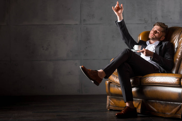 Young businessman in suit sitting on leather sofa holding coffee cup, full length - Foto, Bild