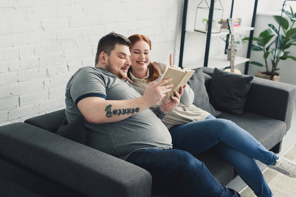 Smiling overweight boyfriend and girlfriend reading book on sofa at home - Zdjęcie, obraz