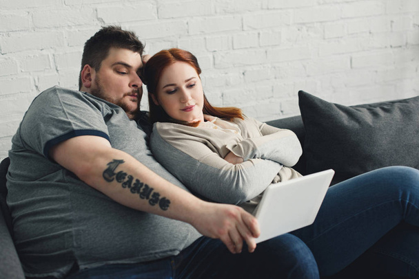boyfriend and girlfriend lying on sofa and watching something on tablet at home - Фото, изображение