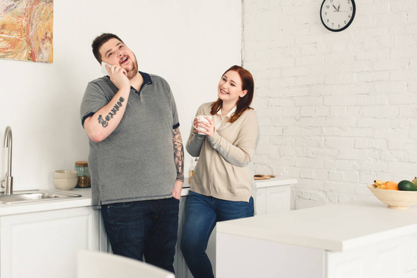happy boyfriend talking by smartphone and girlfriend holding cup of coffee in kitchen - Foto, imagen