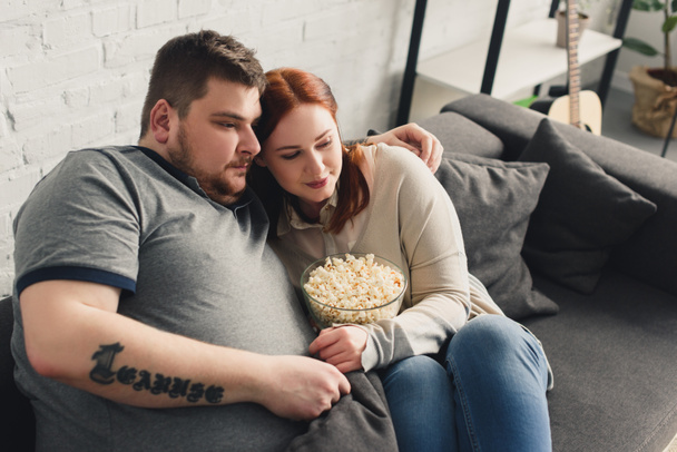 boyfriend and girlfriend hugging on sofa with popcorn and watching movie at home - Фото, изображение