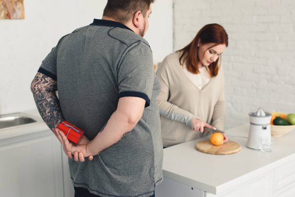 boyfriend hiding present while girlfriend cutting orange in kitchen - Photo, Image