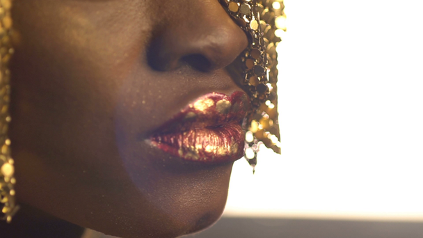 Lábios vermelhos brilhantes cobertos com vasos de bronze dourado no rosto da menina africana encantadora vestindo jóias de ouro em sua cabeça. Close-up
. - Filmagem, Vídeo