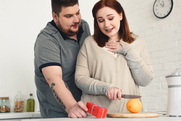 overweight boyfriend presenting gift to girlfriend while she cutting orange at home - Photo, Image