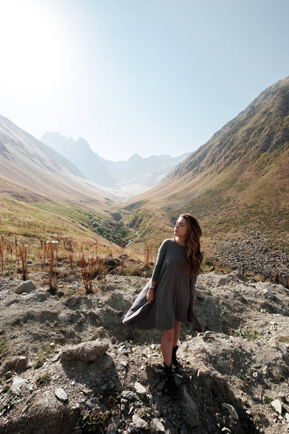Beautiful young woman in a long dress is standing in the valley of the mountains - Фото, изображение